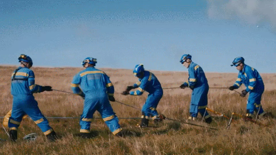 Five coastguard rescue officers in blue overalls stand pulling a rope