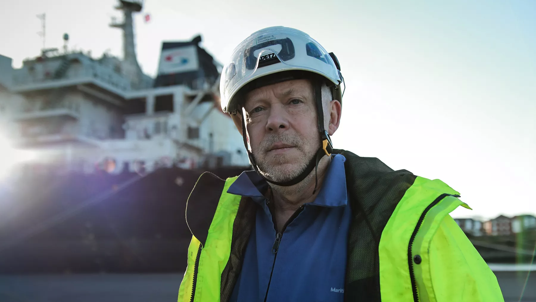A male surveyor in a high-vis jacket looks to the camera. In the background a large ship is blurred from view.