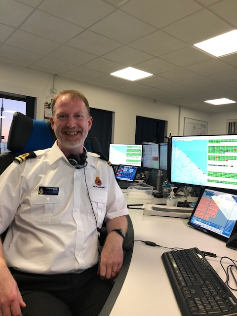 Smiling man with beard in HM Coastguard uniform looking at camera with computer screens behind