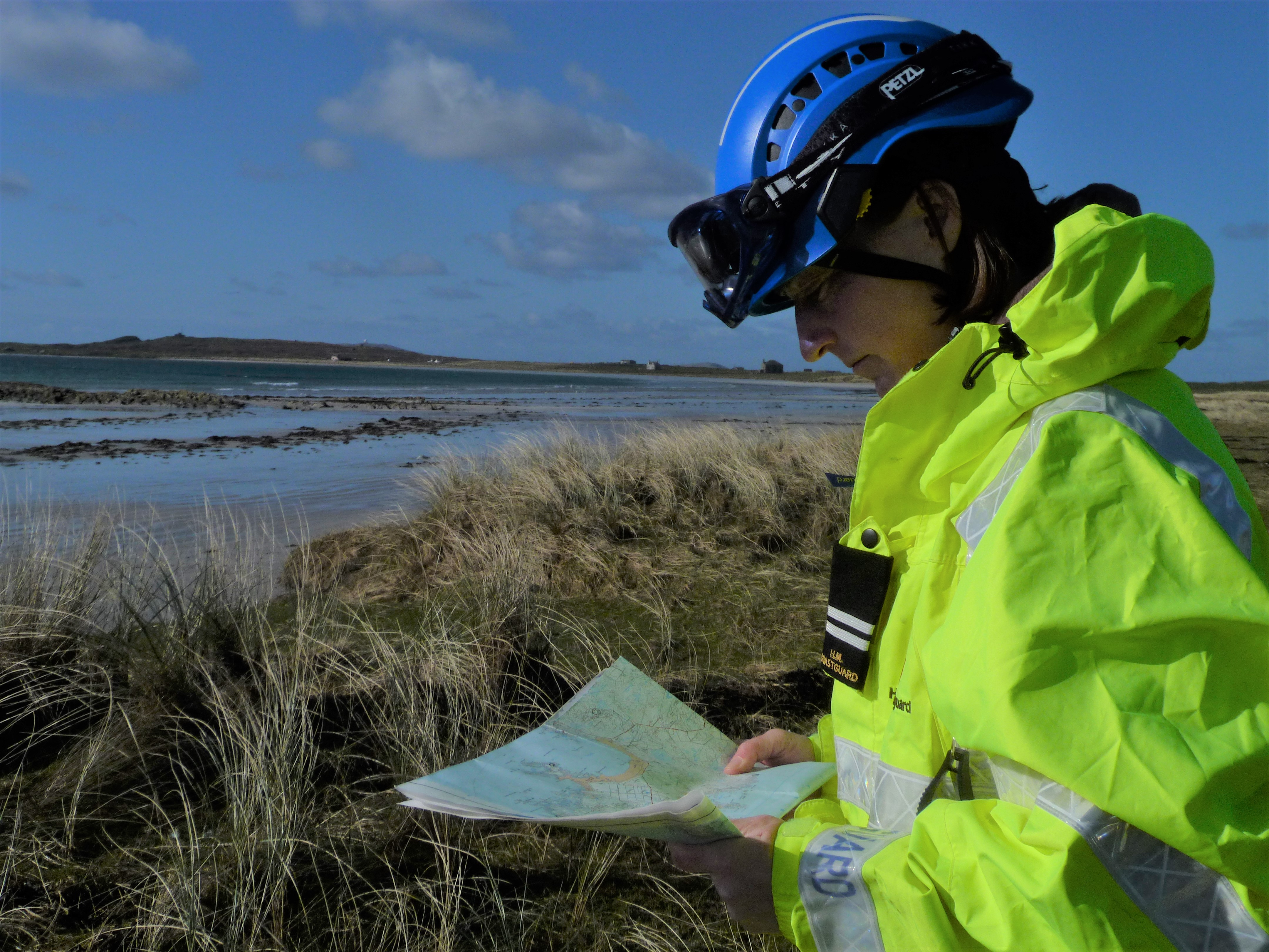 Side view of female Coastguard in PPE and high vis with map in hand and coastal environment in background
