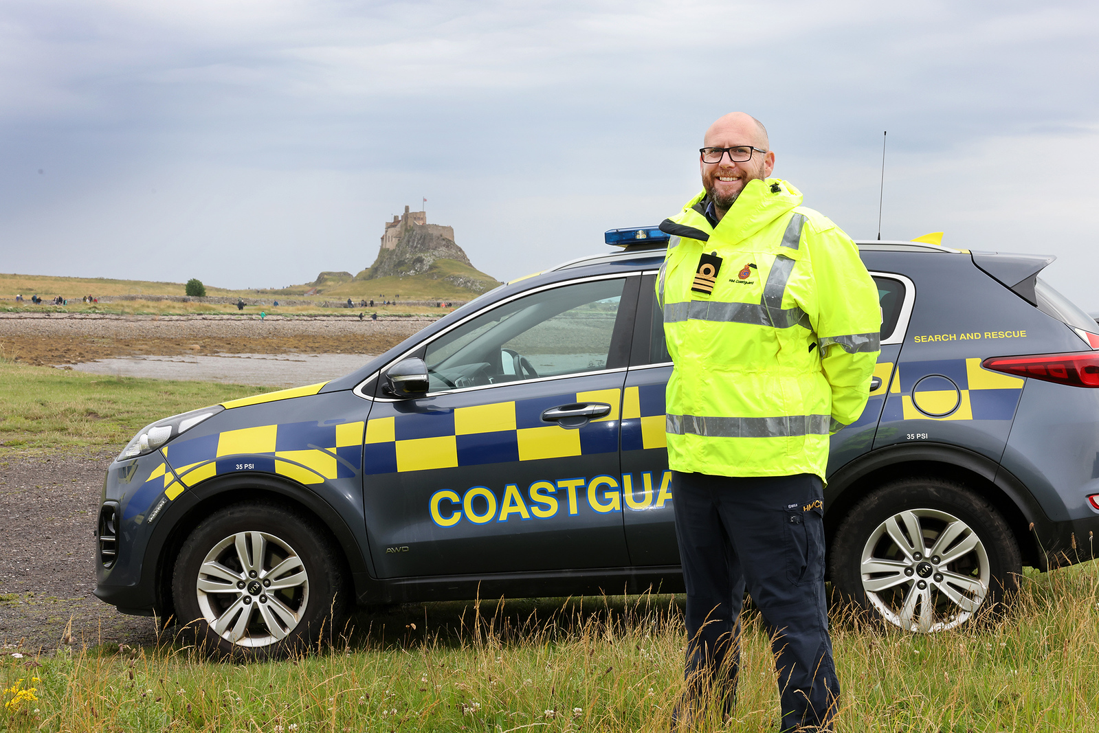 Smiling Coastguard in high-vis jacket in front of Coastguard liveried vehicle and Holy Island in background