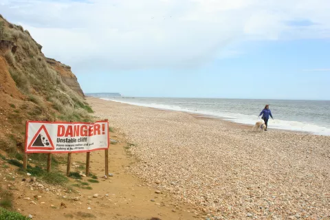 A walker on a beach near a cliff hazard warning sign.