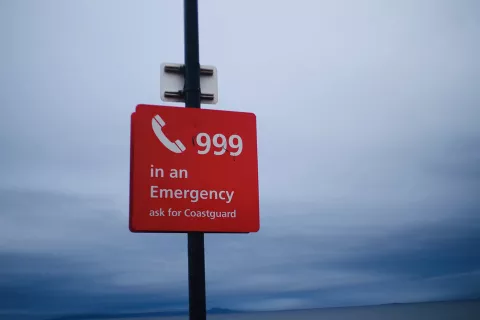 Red sign with 'call 999 in an emergency, ask for the Coastguard' with grey sky backdrop