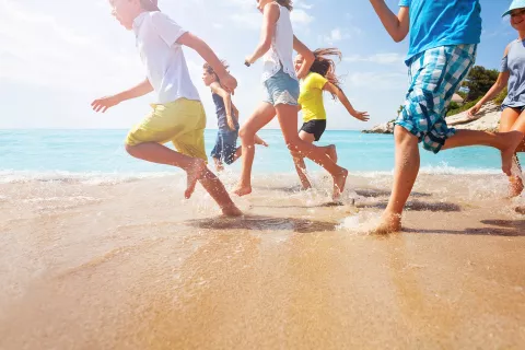 Picture of six children running on sand, focus on legs, near water's edge on sunny day