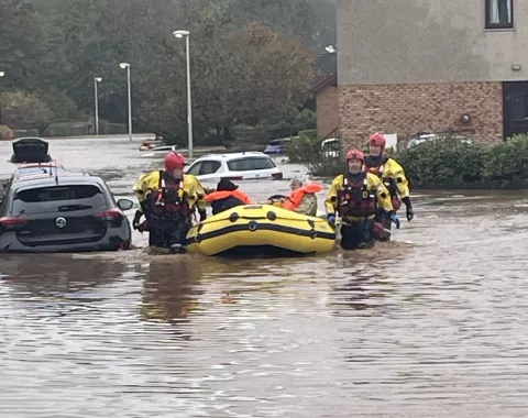 Four men in high vis and with flood rescue equipment wade through waist high water in residential street as they pass a part submerged car