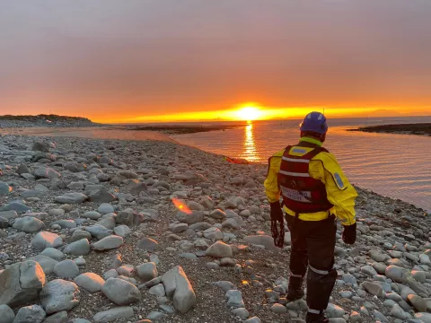 Coastguard Rescue Officer in PPE looking out over water with sun setting on horizon
