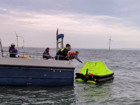 A luminous life raft is collected from the sea by boat crew with offshore wind turbines in the background