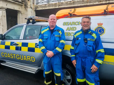 HM Coastguard Fleetwood Coastguard Rescue Team members Steve and Alan stand in blue emergency boiler suits beside the team's Coastguard Rescue Vehicle. 