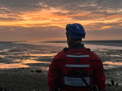 A member of the Southend Coastguard Rescue Team stands on the shoreline amid a golden sunset and low tide. 