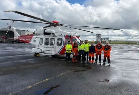 Members of the AW189 helicopter crew at Carlisle, some in orange overalls and others in high-visibility jackets, standing in front of a white and red helicopter. From left to right: Frank Napier (Engineer), Rich Taylor (Winch Operator), Michael Walker (Engineer), Daniel Perry (Engineer), Cathy Freeman-Brown (Winch-Paramedic), Simon White (Captain), Simon Price (Co-Pilot).
