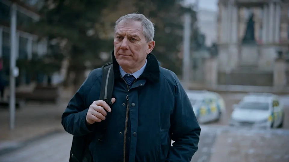 A man holding a backback walks along a city street. In the distance, two parked police cars can just be made out.