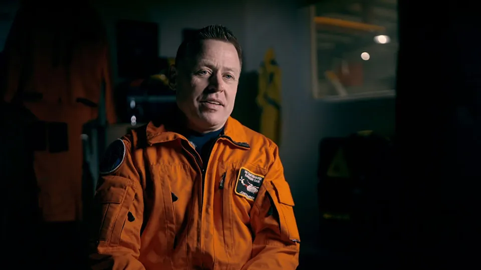 A man in an orange flight suit sits facing the camera. He is a winch paramedic for HM Coastguard