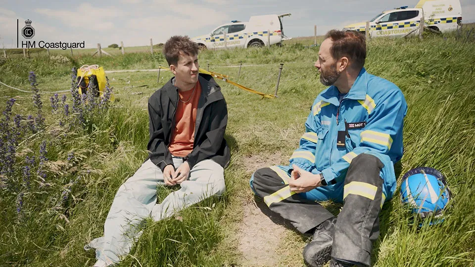 A young man sits on grass next to a coastguard rescue officer. In the distance, two coastguard rescue vehicles are parked.