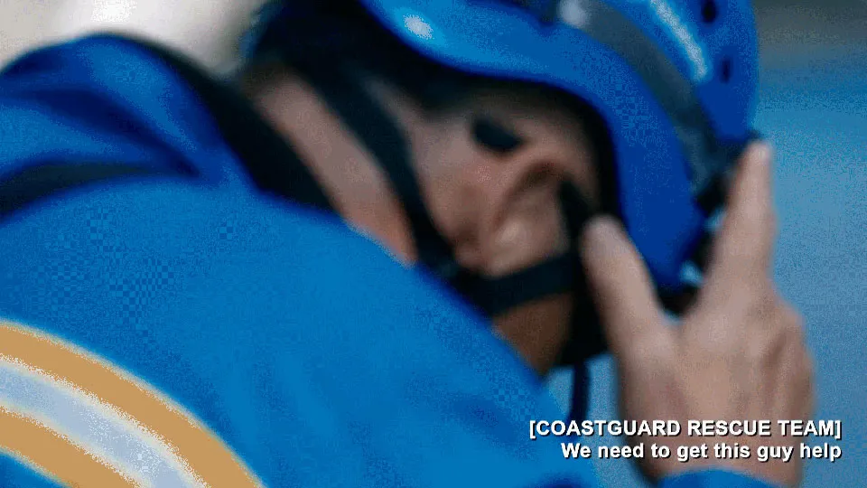 A blurred view of a male coastguard rescue officer wearing a blue overalls and hard hat. He is looking downwards with his hand on his head.