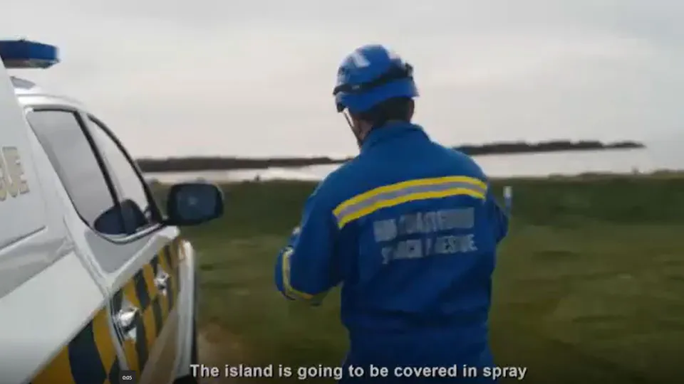 A coastguard rescue officer is wearing blue overalls and is stood next to a coastguard rescue vehicle