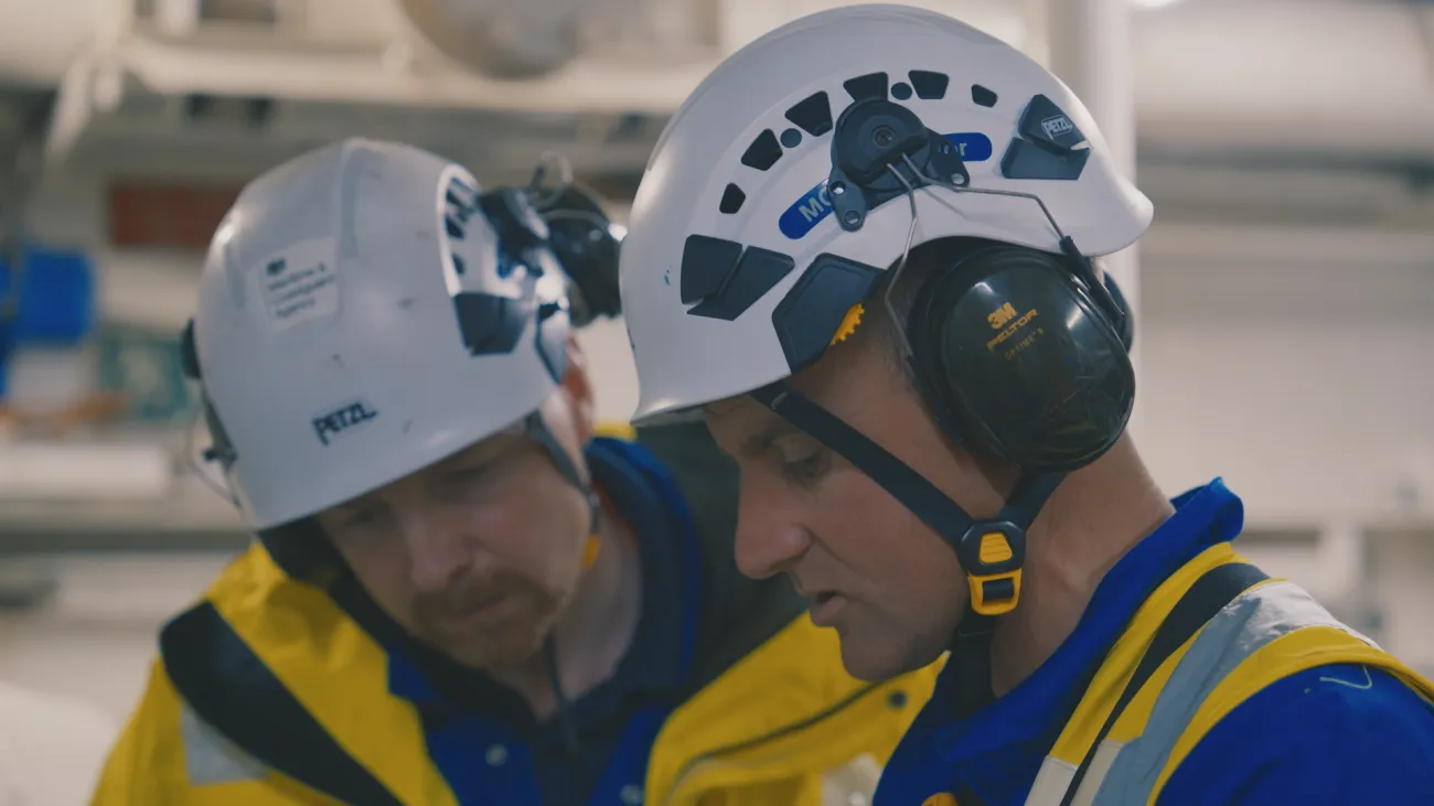 Two male surveyors wearing white helmets are looking down at something off-screen