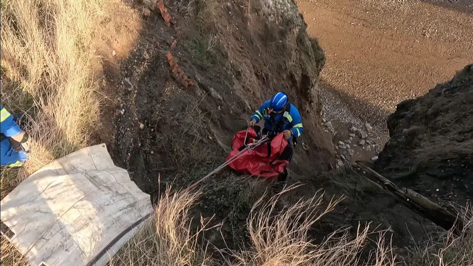 View looking down a cliff. A male coastguard rescue officer can be seen halfway down the cliff face, performing a cliff rescue