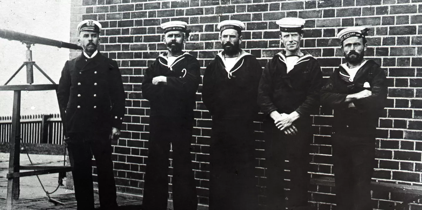 Historic black and white photograph of five coastguard men in uniform stood in front of a brick wall looking towards the camera