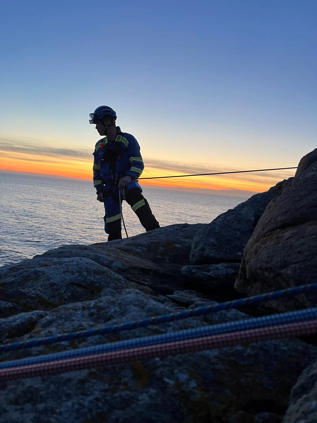 Coastguard rescue officer stands on edge of cliff secure in his rope harness looking down below