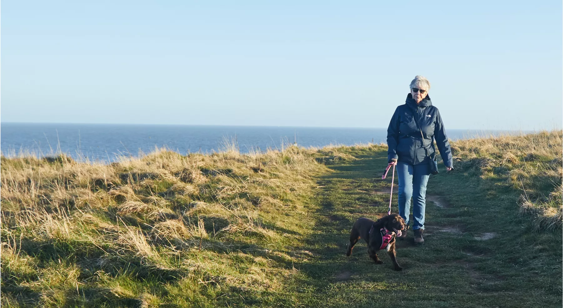 Woman walking her dog along a coastal path with the sea and horizon behind