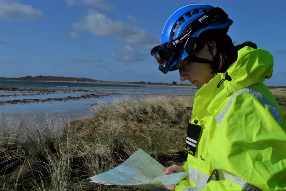Side view of woman in high-vis Coastguard uniform and PPE with map in hand and coastal environment in background
