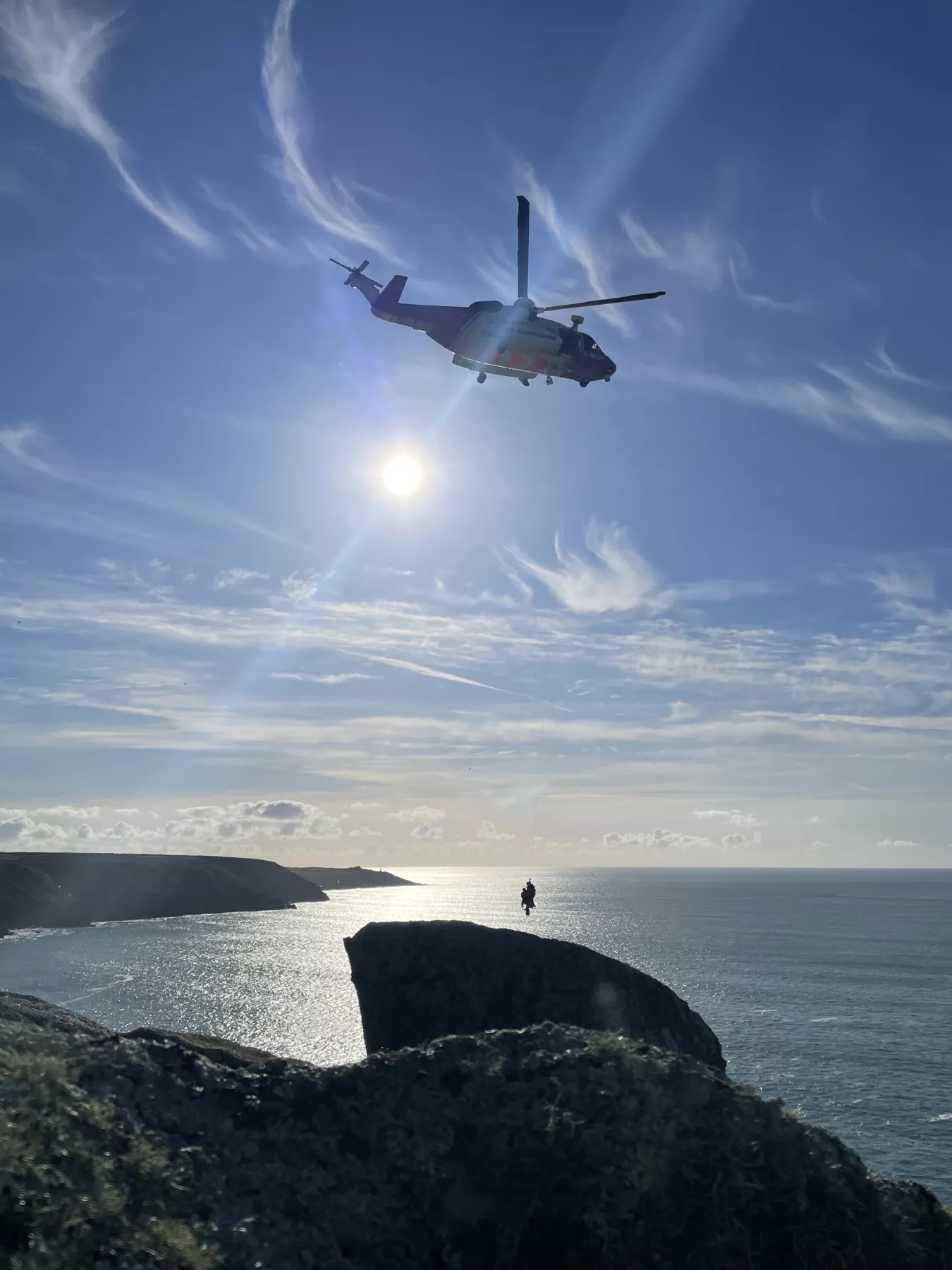 Coastguard helicopter and winch paramedic on the wire over cliff with blue sea and sky