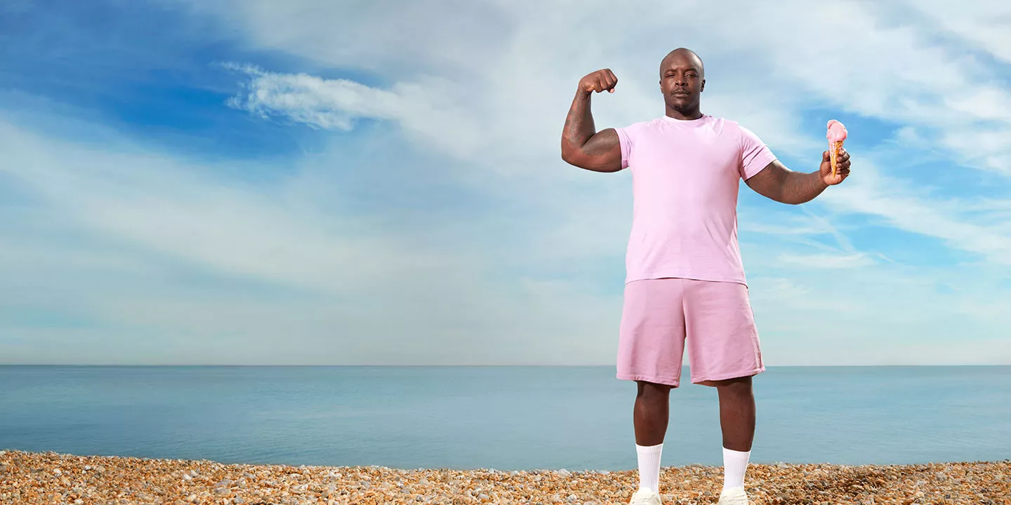 Adebayo Akinfenwa stands on a pebbled beach with blue skies. In one hand he is holding an ice cream, while the other is flexing.