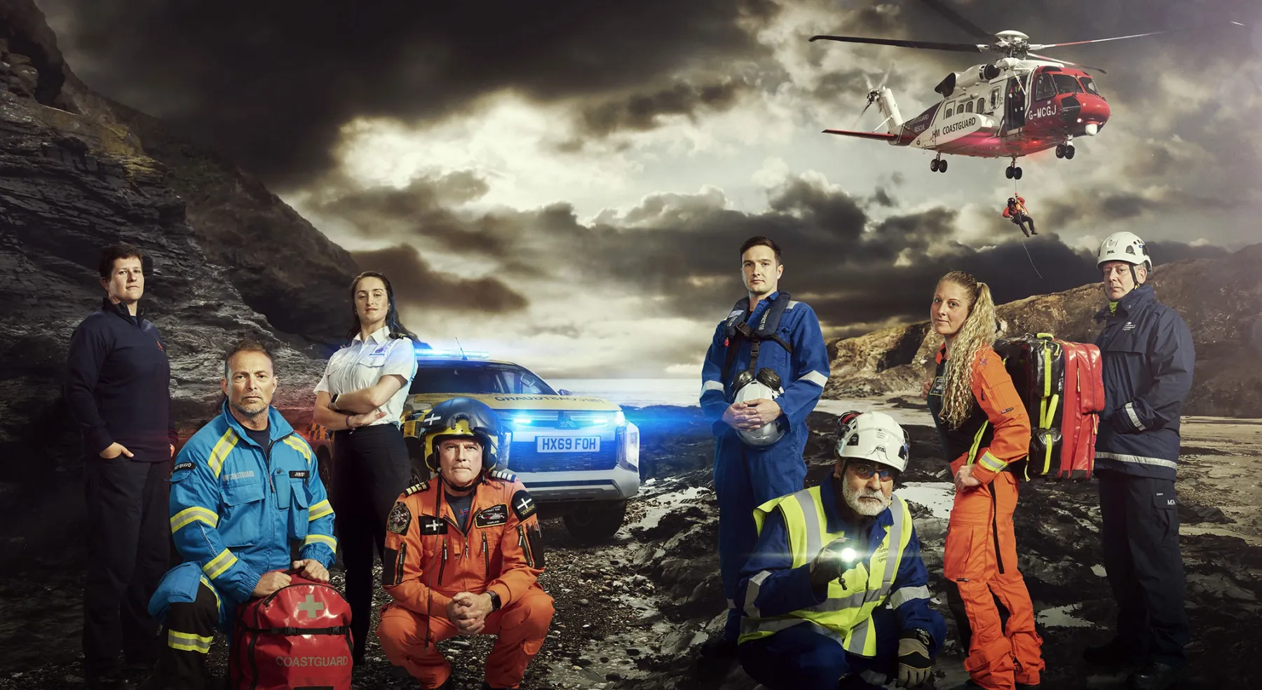 A group of Coastguards in various uniforms stand on a grey, rocky landscape. Above, a Coastguard helicopter is hovering.