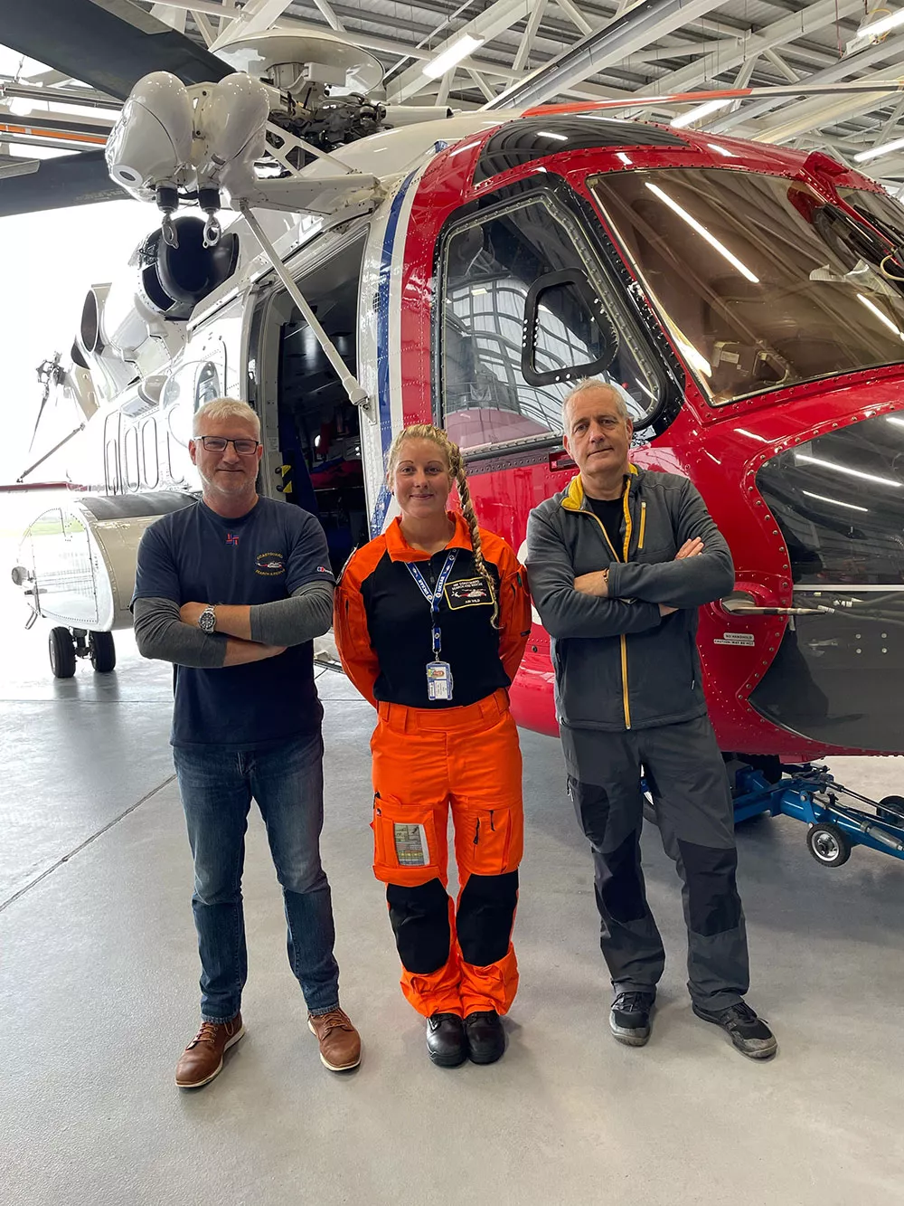 Two men and a woman stand in front of a HM Coastguard helicopter. The woman is dressed in orange winch paramedic overalls.