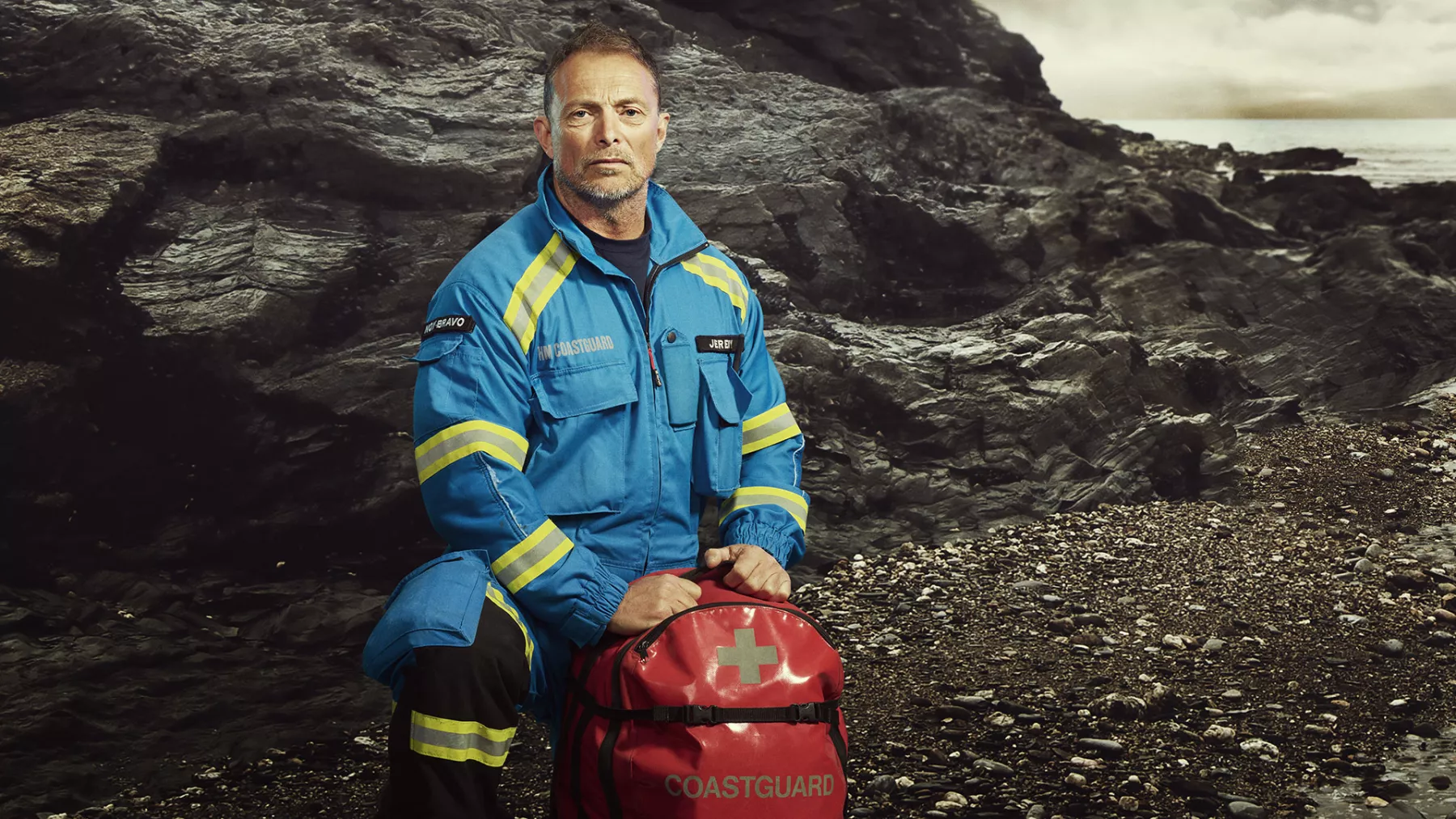 A male Coastguard officer kneels on a grey, rocky landscape. He is wearing his blue and yellow coveralls and has a red first aid kit in his hands.