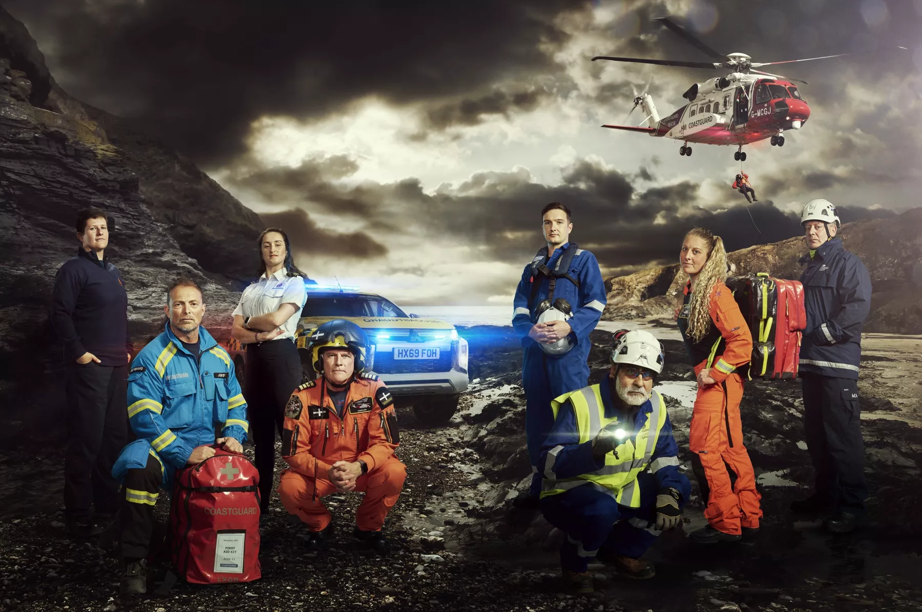 A group of Coastguards in various uniforms stand on a grey, rocky landscape. Above, a Coastguard helicopter is hovering.