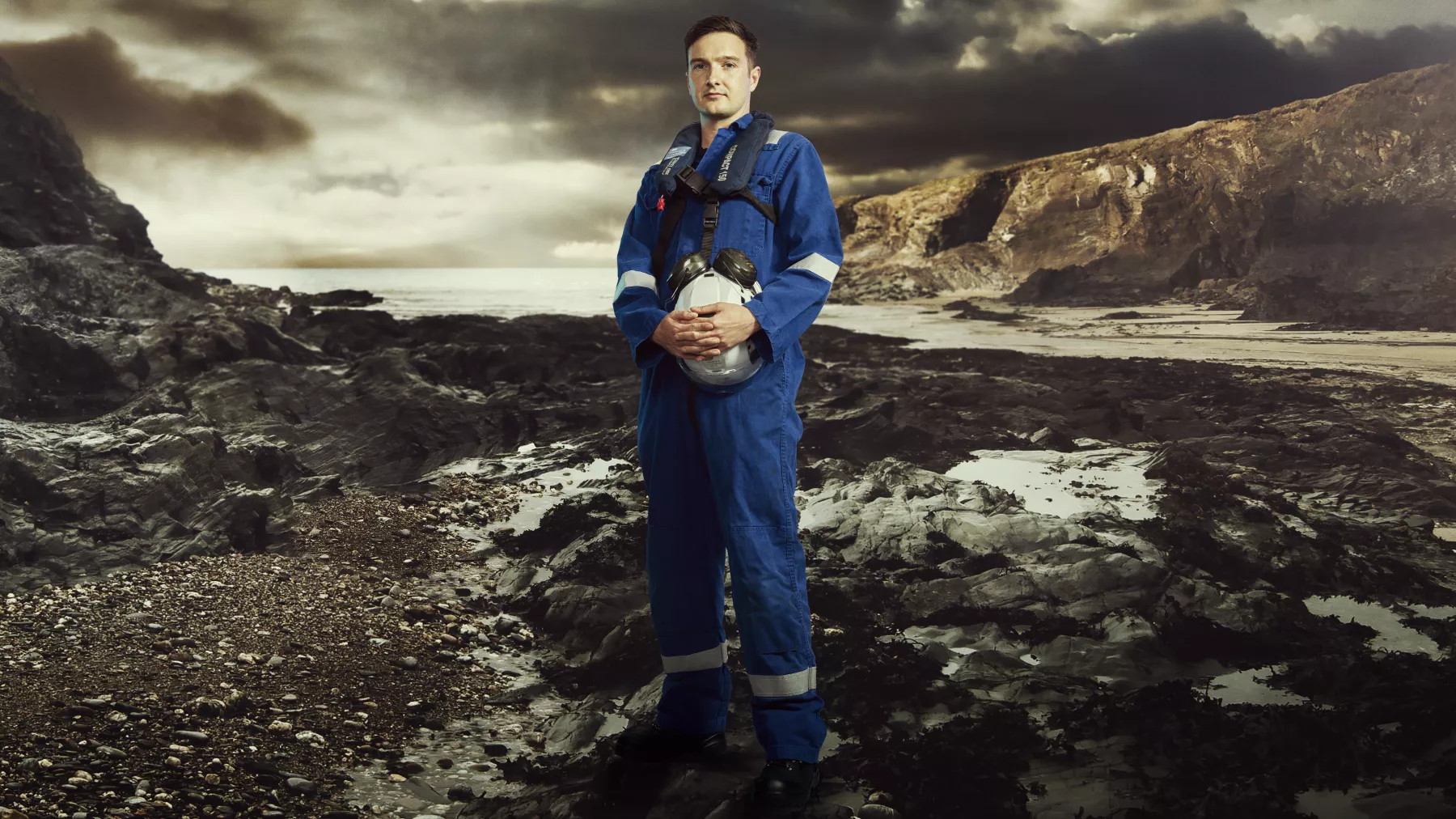 Man in blue coveralls stood on rocky, stormy landscape with white helmet in hands