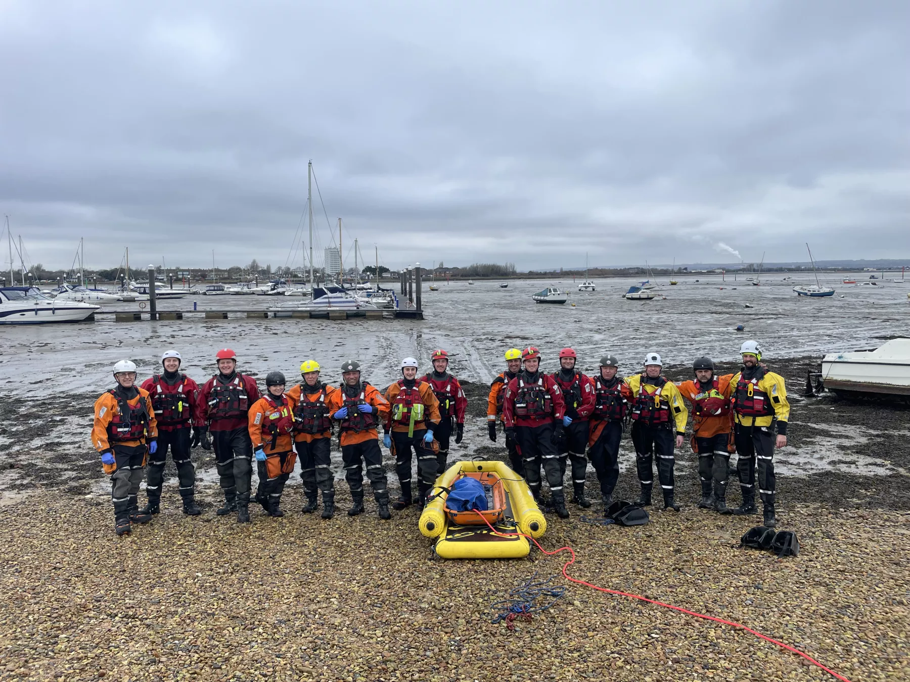 Members of HM Coastguard and Hampshire and Isle of Wight Fire and Rescue Service smiling for a group photo amidst sand and mud in Eastney, Portsmouth, equipped with mud rescue kit