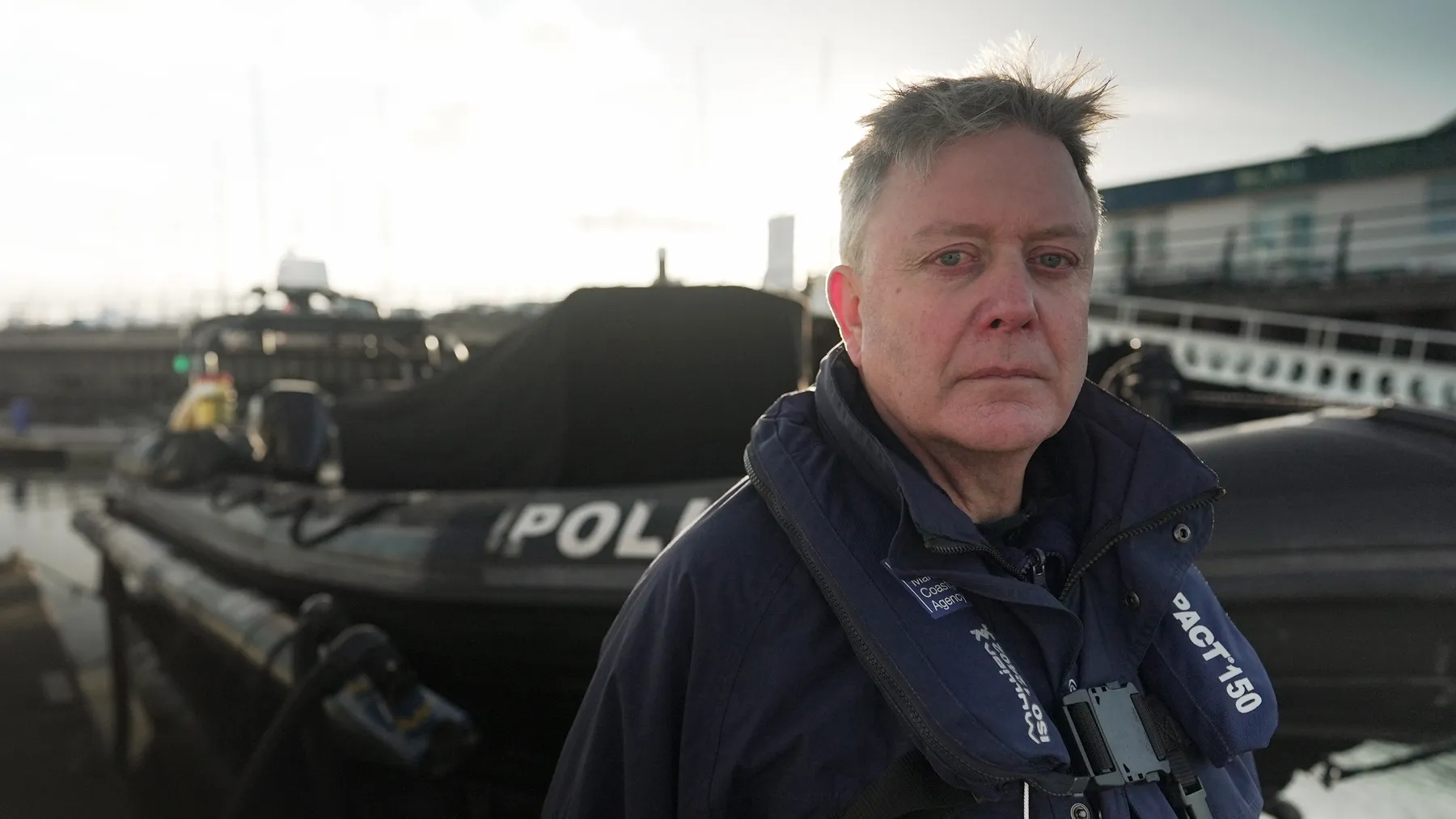 A male senior investigator stands in a harbour wearing a personal floatation device. In the background there is a black police boat