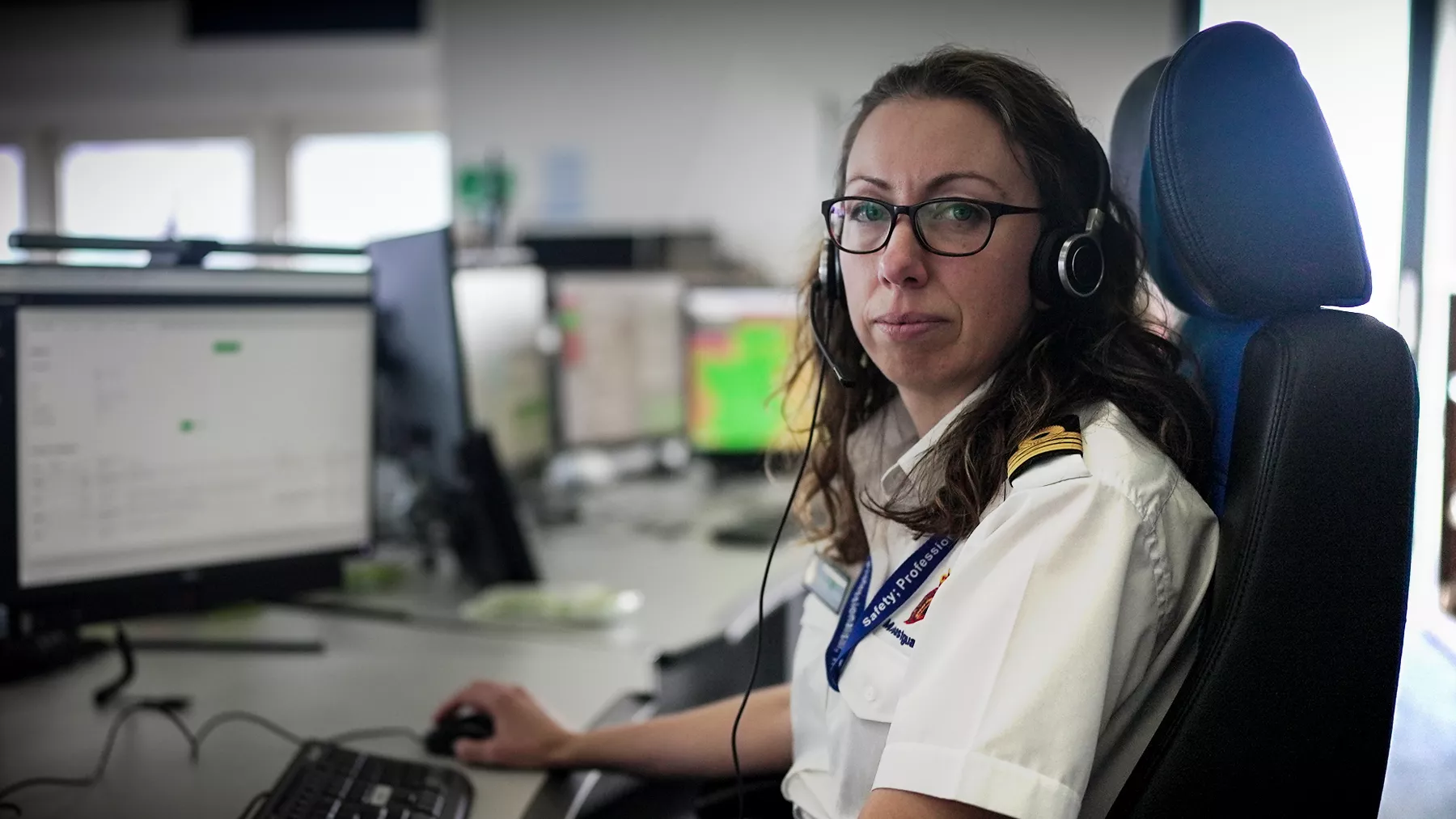 A female maritime operations officer sits facing the camera. She is wearing an official white shirt with the HM Coastguard logo and epaulettes.