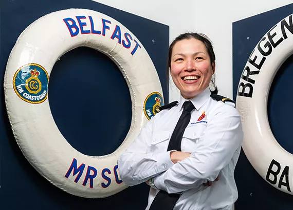 A female Maritime Operations Officer in uniform stands with her arms crossed. Behind, a white lifering hangs on the wall with the words 'Belfast MRSC' written on it