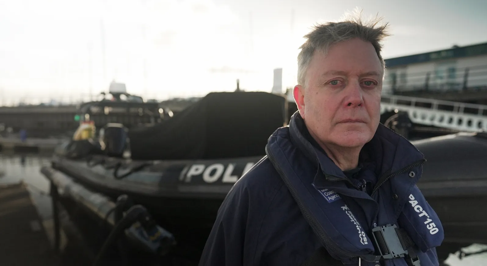 A male Regulatory Compliance Investigator looks at the camera while stood in a small harbour. He is wearing a personal floatation device.