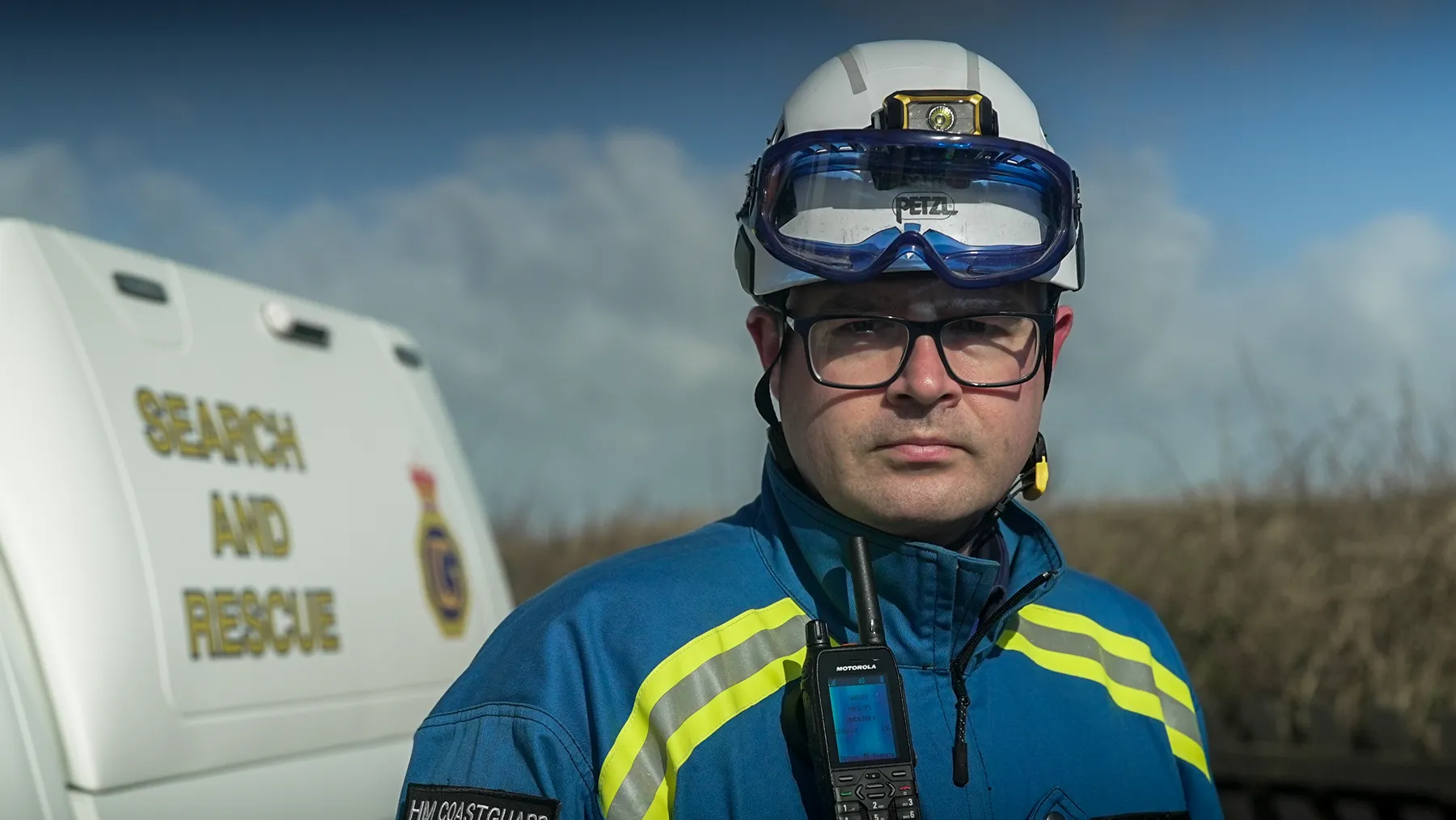 A male coastguard rescue officer stands in front of a coastguard rescue vehicle