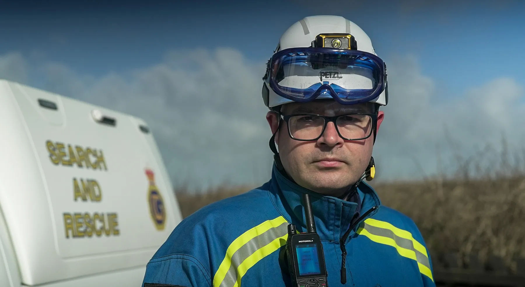 A male coastguard rescue officer wearing blue overalls and a white hard hat stands in front of a coastguard rescue vehicle, looking directly at the camera.