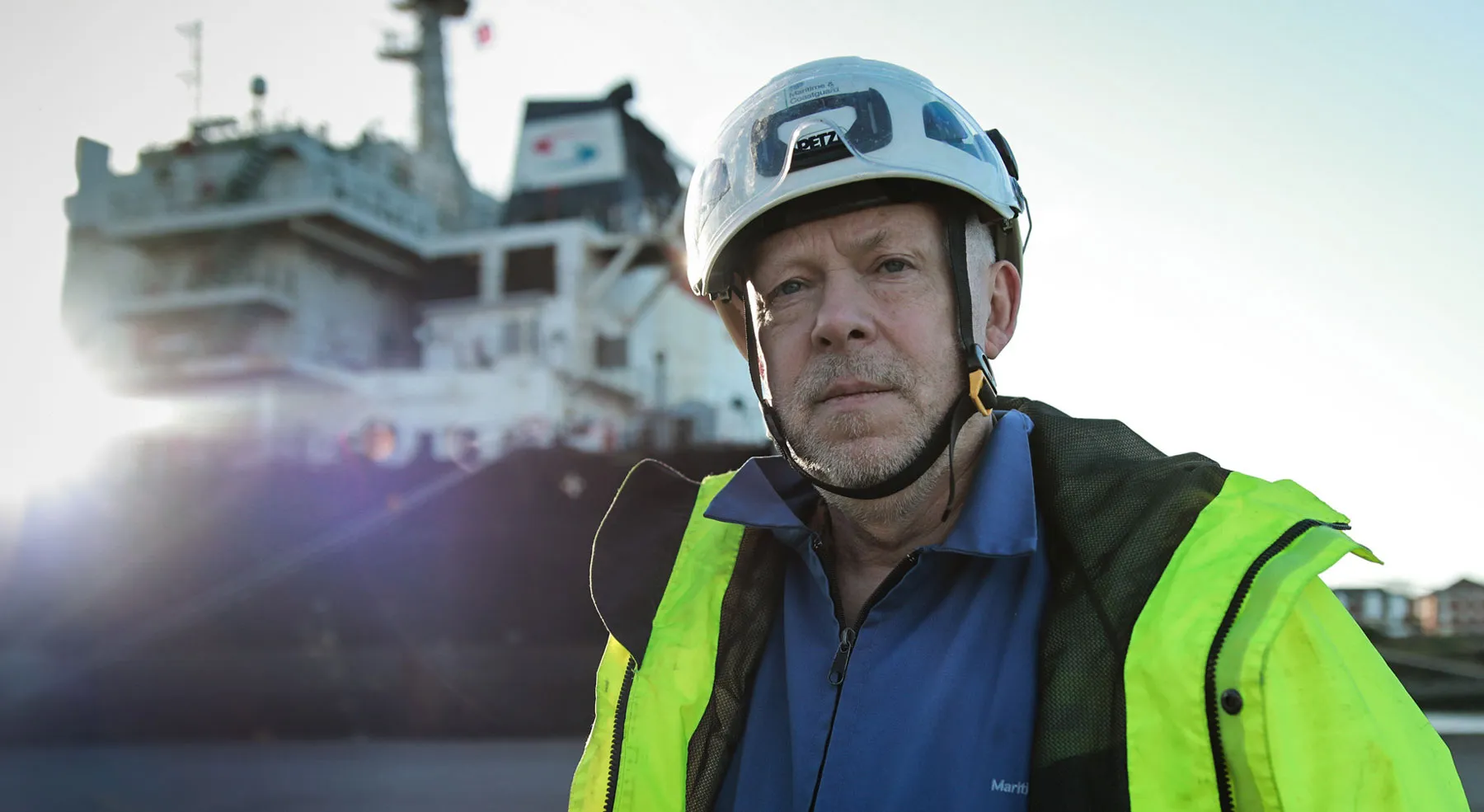 A male surveyor wearing a high visibility jacket and helmet looks at the camera. In the background, a blurred silhouette of a ship can be made out.