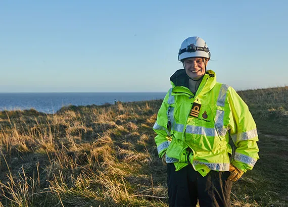 A female Senior Coastal Operations Officer stands facing the camera. In the distance, there are blue skies and seas