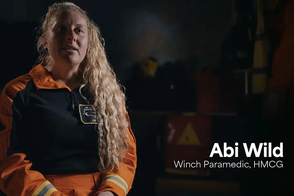 A femal winch paramedic dressed in orange overalls sits against a dark background