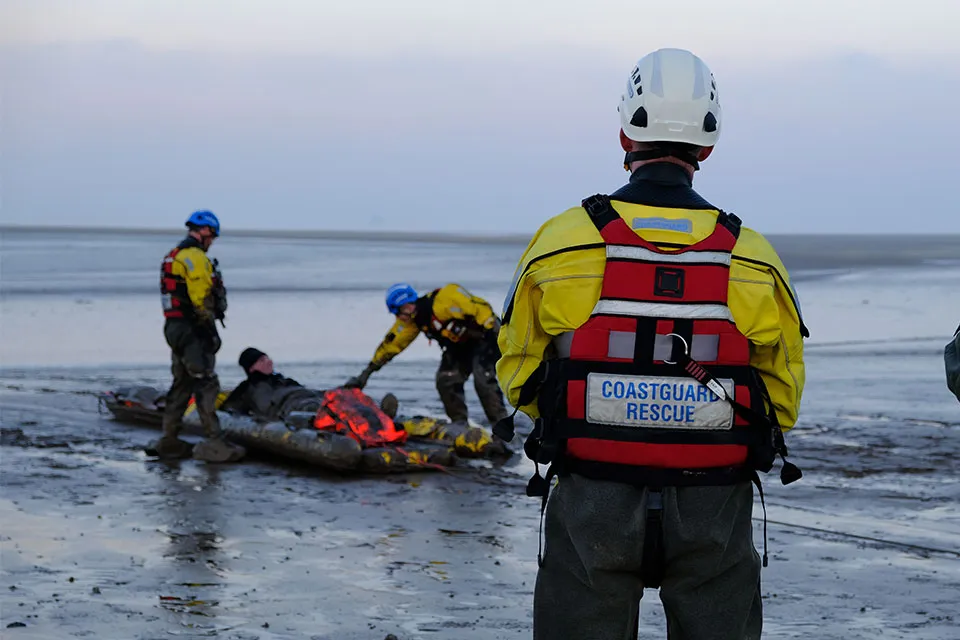 A senior coastal operations officer stands facing away from the camera. In the distance, there are coastguards performing a mud rescue.