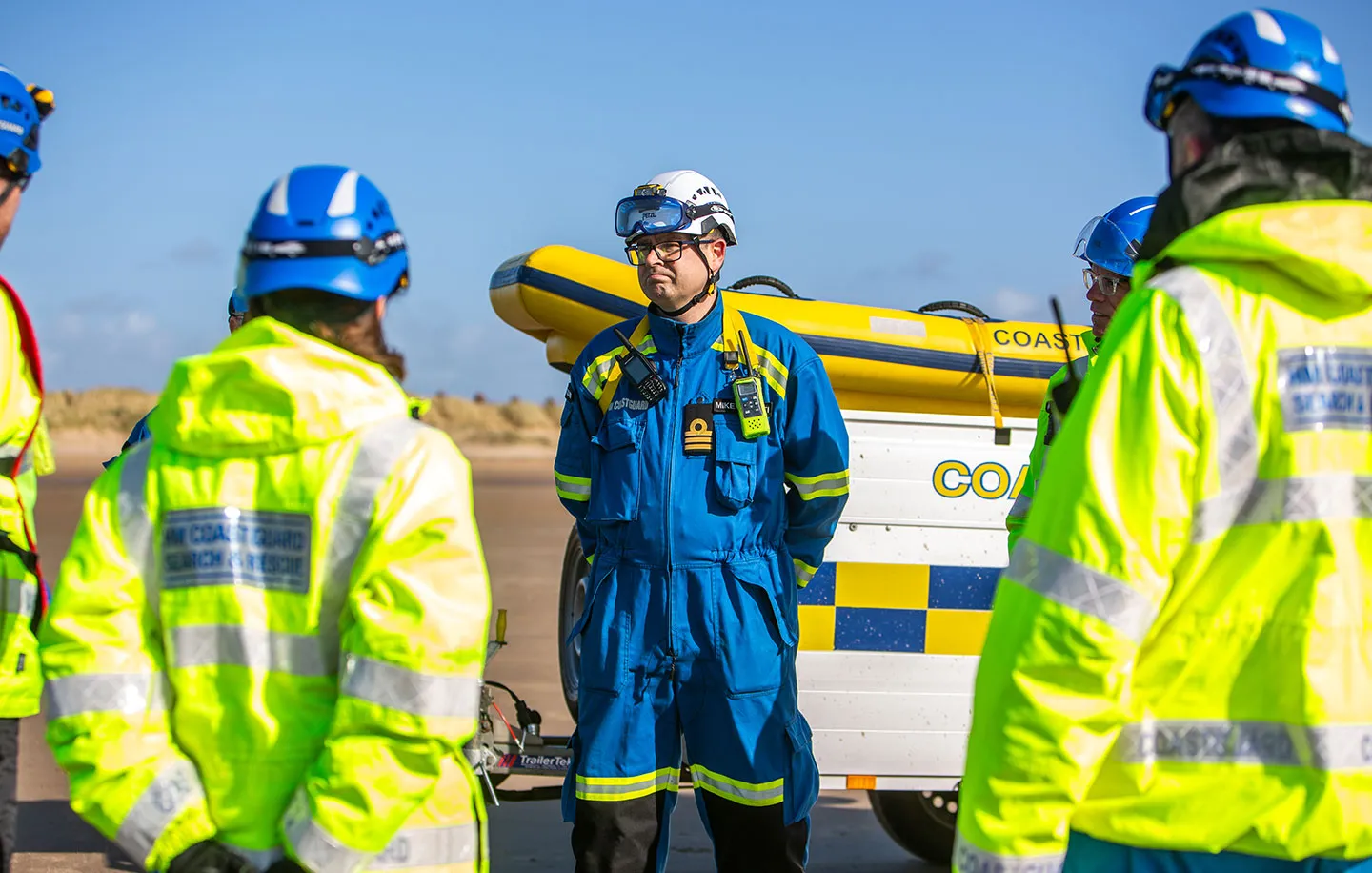A male senior coastal operations officer stands in blue overalls. Beside them are coastguard rescue officers dressed in high-visibility jackets.