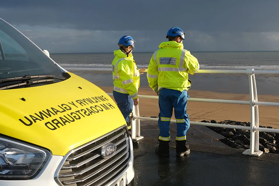 HM Coastguard search and rescue officers stand in front of a coastguard rescue vehicle on a beach promenade. They are looking out to sea where there are grey clouds and a moody sky
