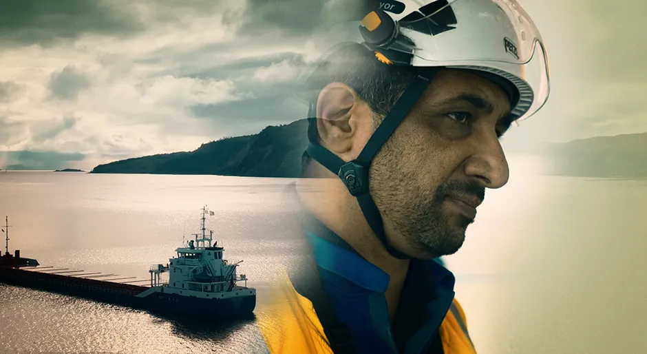 A male surveyor in a high-vis jacket looks to the camera. In the background a large ship is blurred from view.