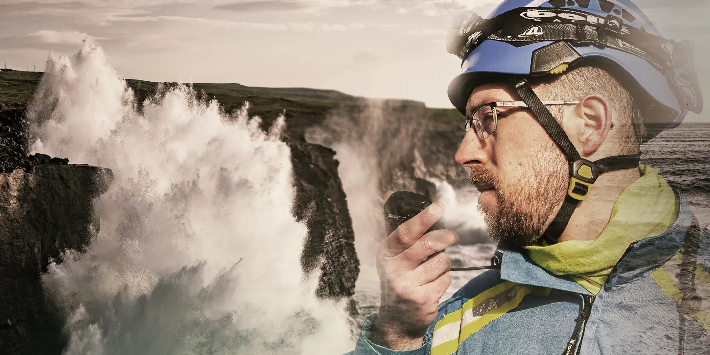 A moody coastline with large waves crashing onto the rocks on the left. Overlaid on the right, a male coastguard rescue officer is speaking into a radio. He is wearing a blue helmet and blue HM Coastguard overalls.