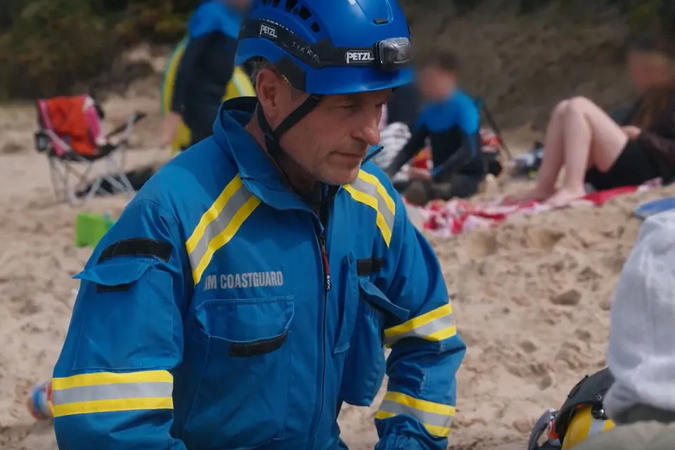 A coastguard rescue officer, in blue HM Coastguard overalls, is on a busy sand beach