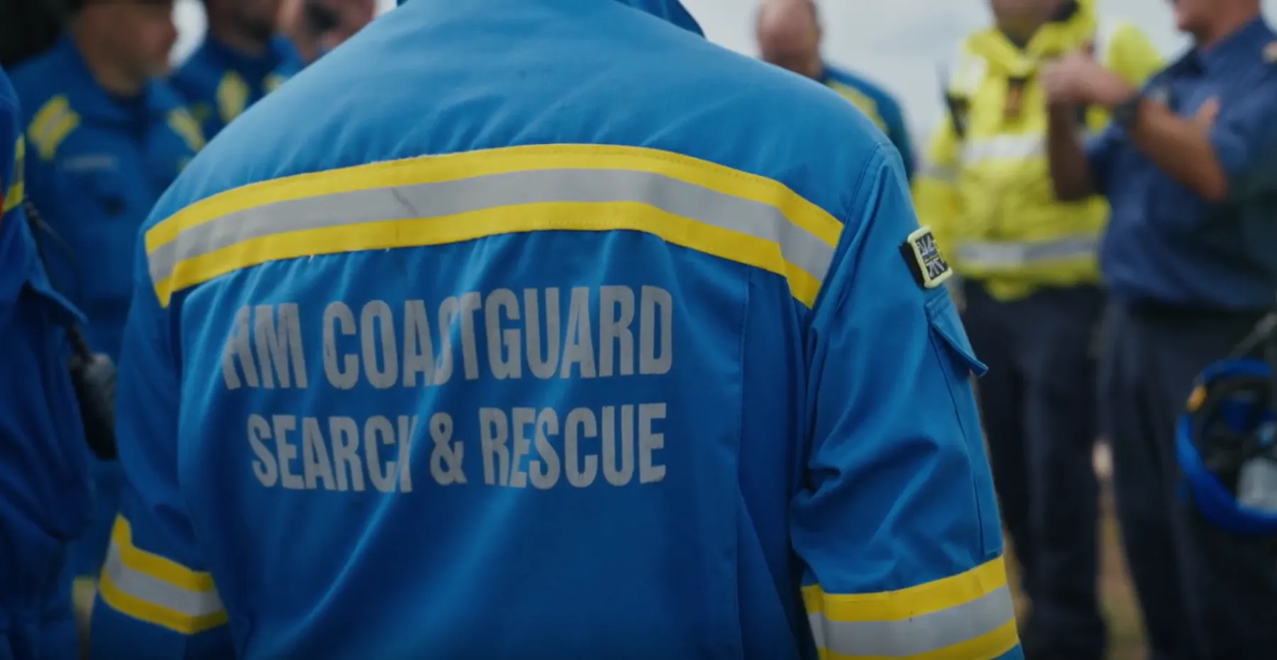 The back of a coastguard rescue officer in blue overalls. On the overalls, text reads 'HM Coastguard search and rescue'