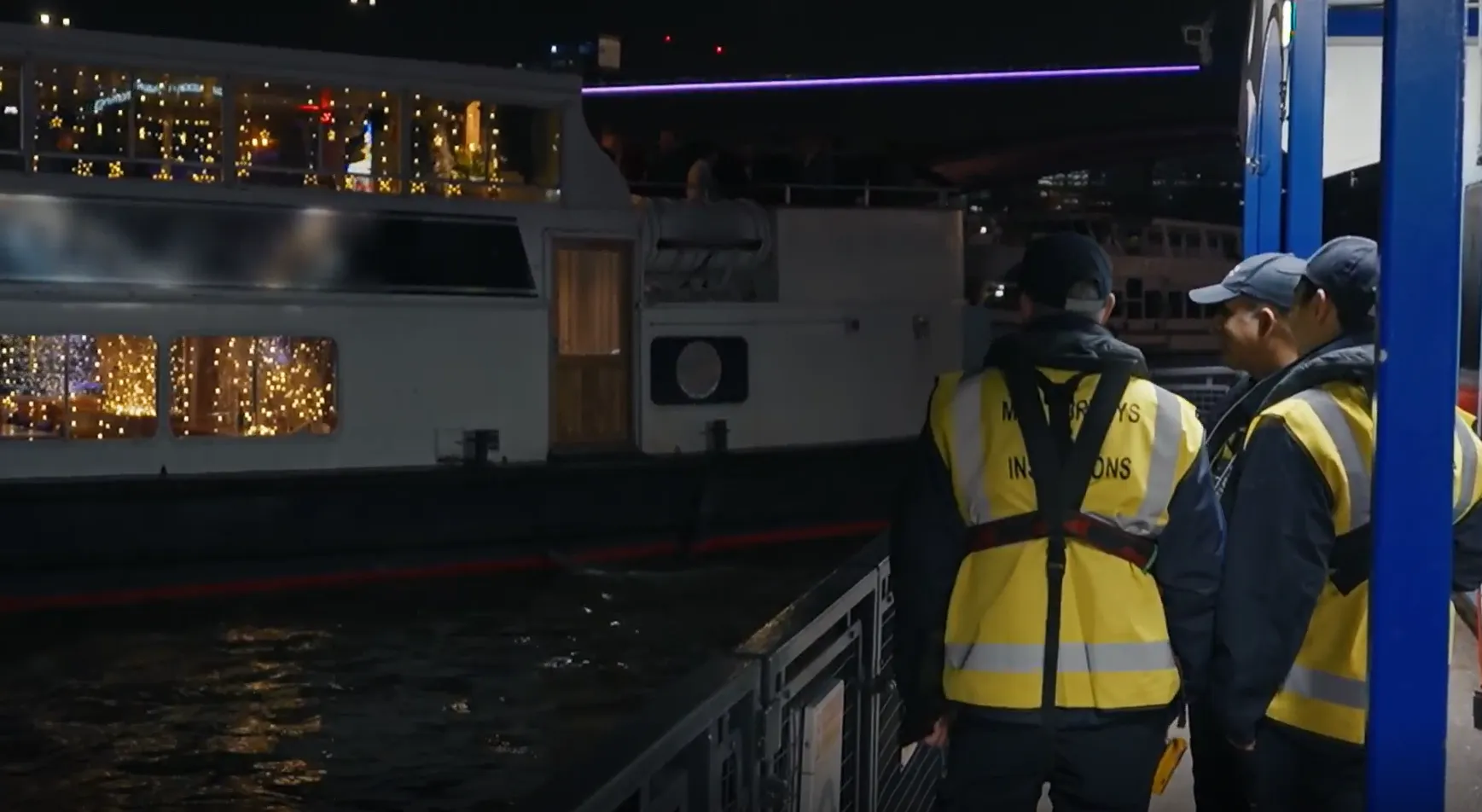 Individuals wearing high visibility jackets with the words 'survey and inspection' on stand looking at a boat adorned with lights